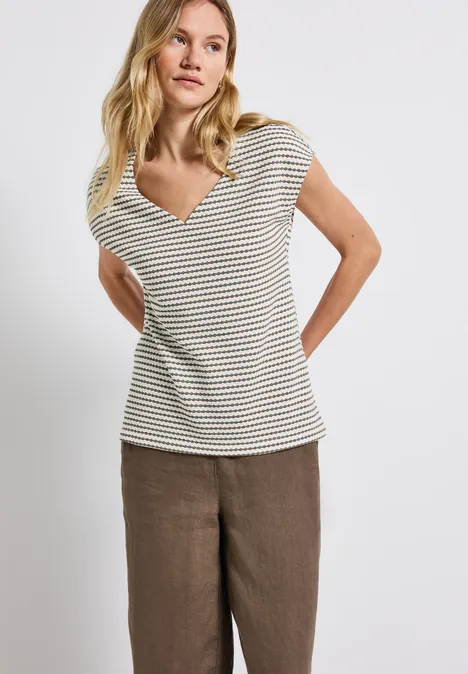 Frau mit gestreiftem T-Shirt und brauner Hose steht vor weißem Hintergrund.